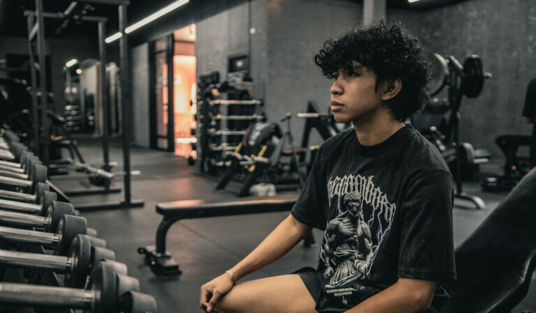a young man sitting on a gym bench in front of a dumbbell rack and looking forward