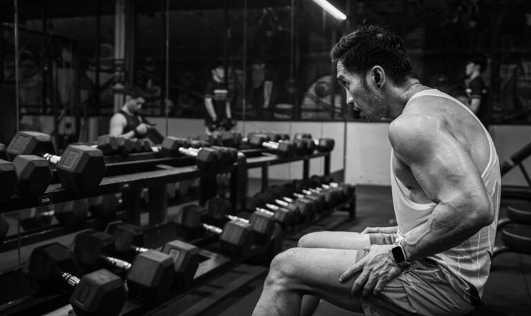 muscular man wearing a tank top sitting on a gym bench in front of a dumbbell rack and a mirror