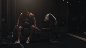 a muscular man wearing a tank top and shorts seated on a workout bench inside a gym
