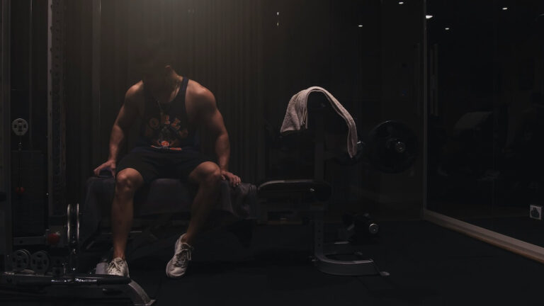 a muscular man wearing a tank top and shorts seated on a workout bench inside a gym
