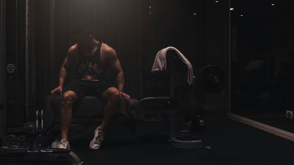 a muscular man wearing a tank top and shorts seated on a workout bench inside a gym