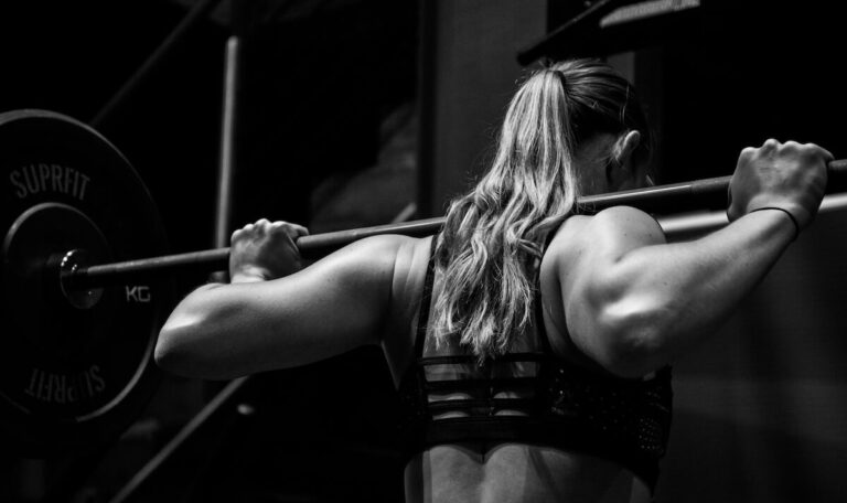 photo of a woman with a barbell placed on her upper back with a gym background